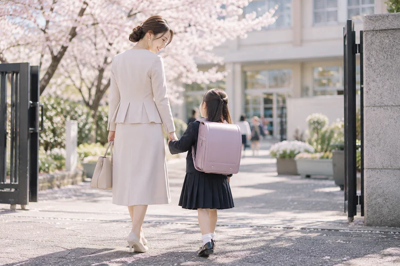入園式・入学式に向かう桜並木の道で、ミモレ丈のベージュセットアップを着た母親がランドセルを背負った子どもと手をつないで歩いている後ろ姿