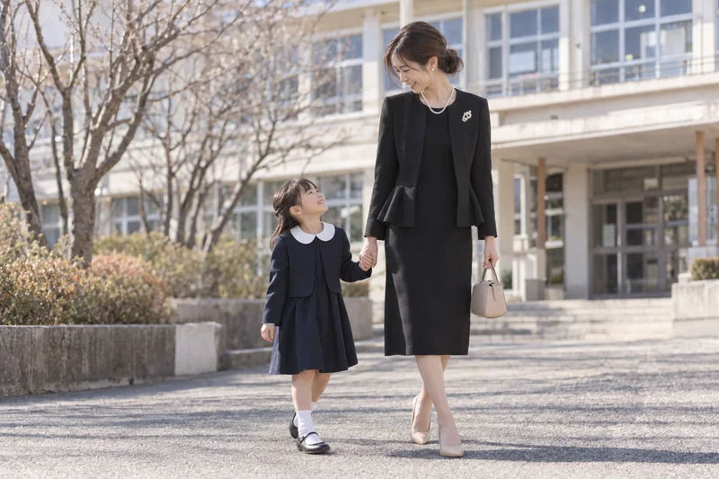 卒業式・入学式の日、小学校の校舎前で娘と手をつないで歩くママ。黒のペプラムジャケットとワンピースにパールネックレスとブローチを合わせた上品な黒ジャケットコーデ