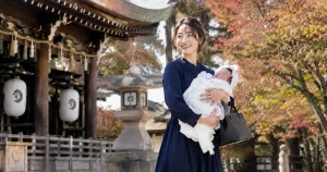 紅葉の中、赤ちゃんを抱っこして微笑む紺色ワンピース姿のママ｜秋のお宮参り
