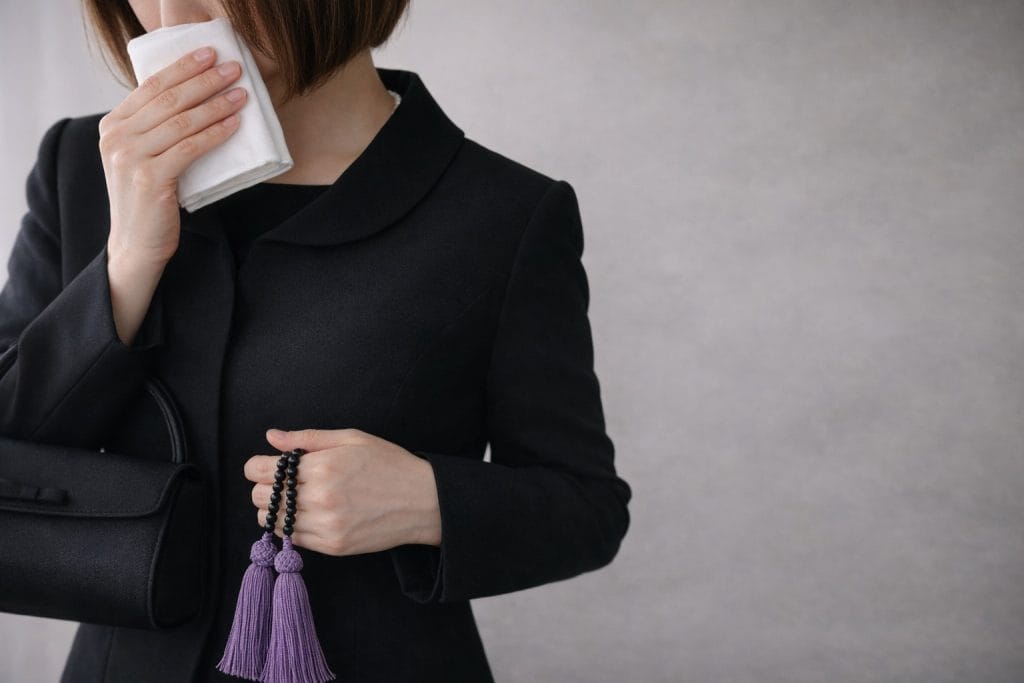 黒の喪服姿の女性が白いハンカチを顔に当てている|数珠を持ったお葬式参列シーン