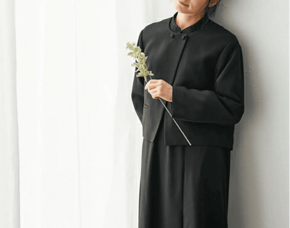 黒のアンサンブル喪服を着た女性が白い壁の前に立つ上品なフォーマルスタイル